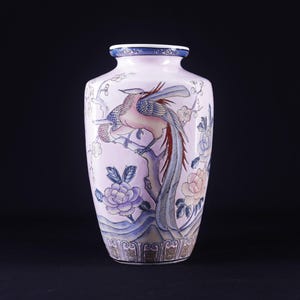May include: A pink ceramic vase with a blue and gold floral design featuring two birds with long tails. The vase has a rounded body and a narrow neck.