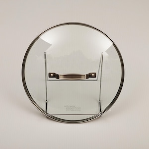 May include: A clear glass pot lid with a stainless steel rim and handle. The lid is resting on a stainless steel stand. The text "KITCHEN essentials" is printed on the glass.