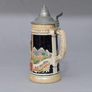 May include: A ceramic beer stein with a dark blue body featuring a relief scene of a mountain landscape, a house, and the text Auf der Alm. It includes a metallic conical lid with a thumb lever and a textured, light-colored handle.