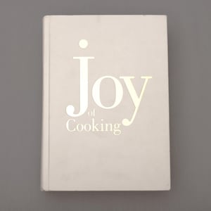 May include: A hardcover cookbook titled "Joy of Cooking" in a light gray color. The title is printed in a lighter, metallic font. The book is closed and centered on a gray background.