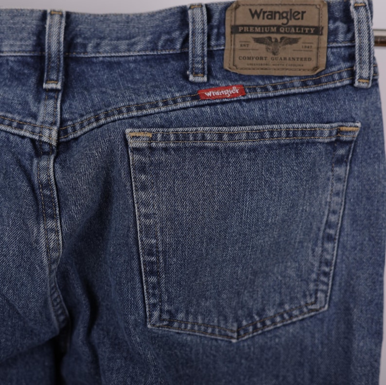 May include: Close-up of blue denim Wrangler jeans. The jeans feature a back pocket, belt loops, and a brown leather patch with the Wrangler logo. A red Wrangler tag is visible. The jeans are made of a durable denim material.