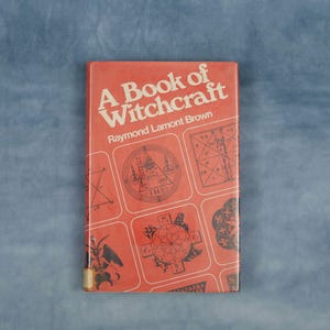 Puede incluir: Un libro antiguo titulado "A Book of Witchcraft" de Raymond Lamont Brown. La portada es de color rojo anaranjado con texto blanco y varios s&iacute;mbolos ocultos en cuadrados blancos. El libro es rectangular y parece estar en buen estado.
