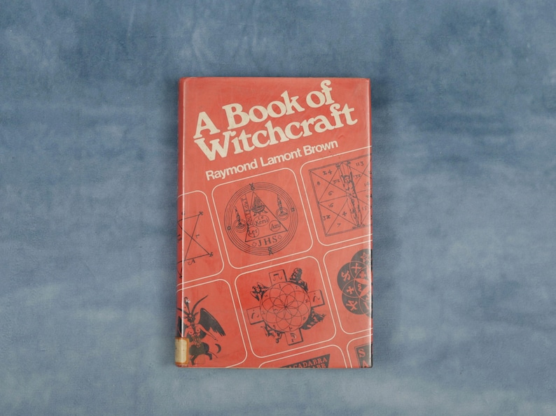 Puede incluir: Un libro antiguo titulado "A Book of Witchcraft" de Raymond Lamont Brown. La portada es de color rojo anaranjado con texto blanco y varios s&iacute;mbolos ocultos en cuadrados blancos. El libro es rectangular y parece estar en buen estado.