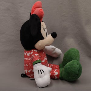 May include: A plush Minnie Mouse doll in a seated position. Minnie has a black head, a red bow, and a red and white patterned outfit. Her gloves are white, and her shoes are green. The doll is on a gray background.