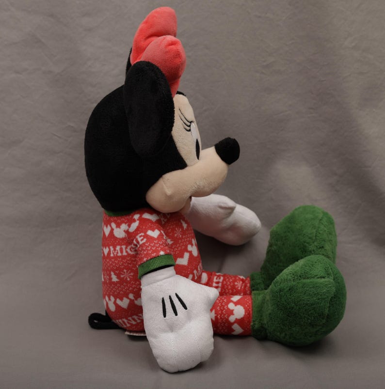 May include: A plush Minnie Mouse doll in a seated position. Minnie has a black head, a red bow, and a red and white patterned outfit. Her gloves are white, and her shoes are green. The doll is on a gray background.