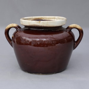 May include: A dark brown ceramic pot with two side handles and a wide, cream-colored rim. The vessel features a glossy, smooth finish on the body and a slightly textured, lighter glaze around the top opening and the handles, creating a contrast.