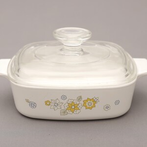 May include: A white square casserole dish with a clear glass lid and handle. The dish features a floral pattern in yellow, blue, and green. The dish has two white handles on either side.