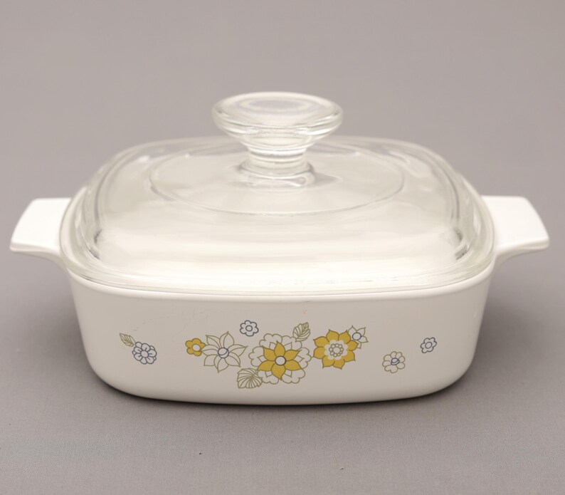 May include: A white square casserole dish with a clear glass lid and handle. The dish features a floral pattern in yellow, blue, and green. The dish has two white handles on either side.