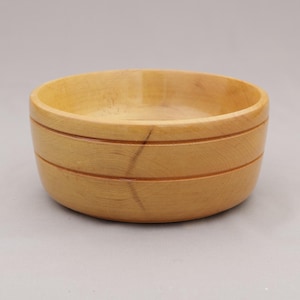 May include: A light-colored wooden bowl with a round shape. The bowl has two horizontal grooves carved around the exterior. The interior of the bowl is smooth and empty. The bowl is made of a light-colored wood.