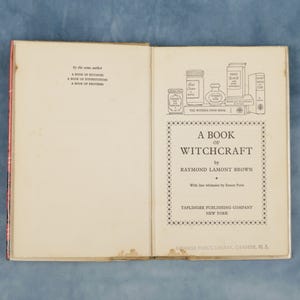 Puede incluir: Libro abierto titulado "A Book of Witchcraft" de Raymond Lamont Brown. La portada presenta ilustraciones de tarros y botellas. Publicado por Taplinger Publishing Company, Nueva York.