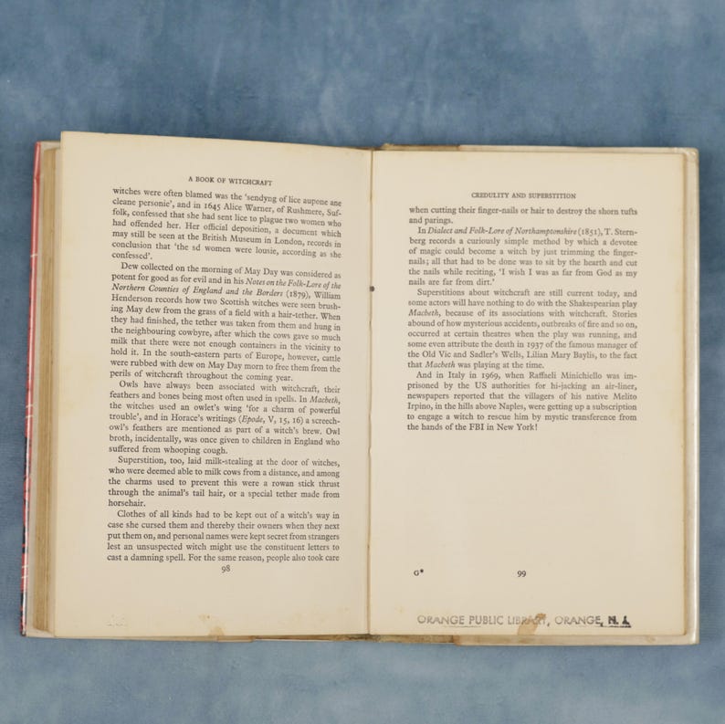 Puede incluir: Libro antiguo abierto con textos sobre brujer&iacute;a y superstici&oacute;n. La p&aacute;gina izquierda detalla creencias hist&oacute;ricas, la derecha pr&aacute;cticas relacionadas. Las p&aacute;ginas 98 y 99 son visibles, con un sello de la Biblioteca P&uacute;blica de Orange, Orange, MA.
