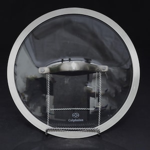 May include: A circular tempered glass lid with a polished stainless steel rim and a matching metal handle. The center of the glass features the Calphalon brand logo printed in white. The lid is designed for cookware and has a clear, transparent surface.