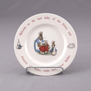 May include: White ceramic plate featuring a Peter Rabbit illustration with a family of rabbits. The plate has text in a circular pattern that reads "Welcome to the new baby of the Prince and Princess of Wales, with love, Peter Rabbit."