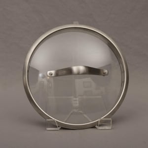May include: A clear glass lid with a stainless steel rim and handle. The lid is round and has a curved glass surface. The handle is a curved bar. The lid is supported by two clear plastic stands.