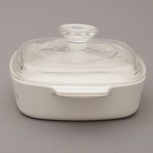 May include: A white square casserole dish with a clear glass lid and a clear glass knob. The dish has two handles and is sitting on a gray surface. The dish is made of ceramic.