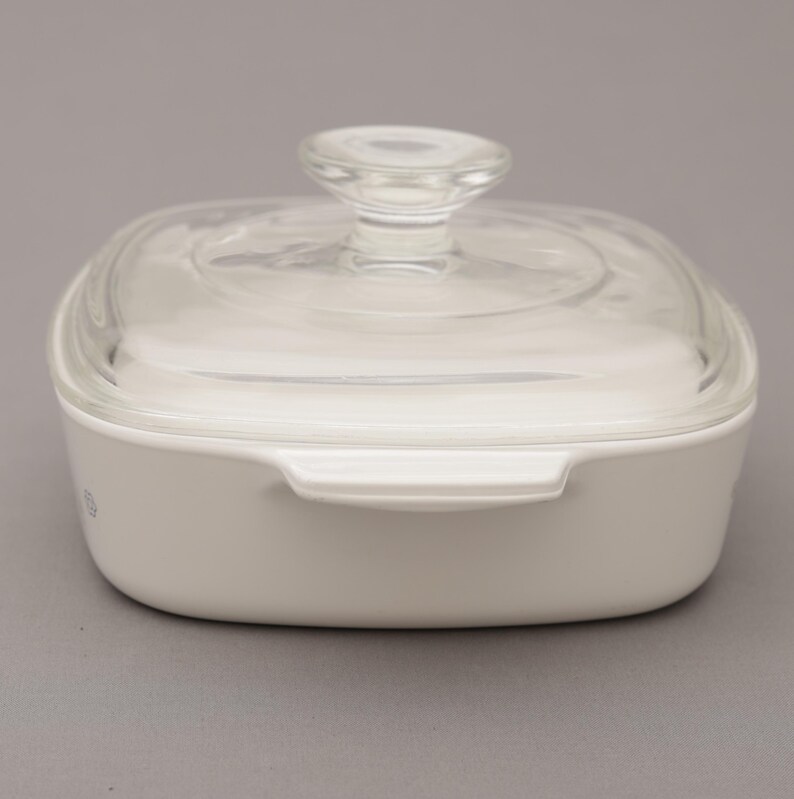 May include: A white square casserole dish with a clear glass lid and a clear glass knob. The dish has two handles and is sitting on a gray surface. The dish is made of ceramic.