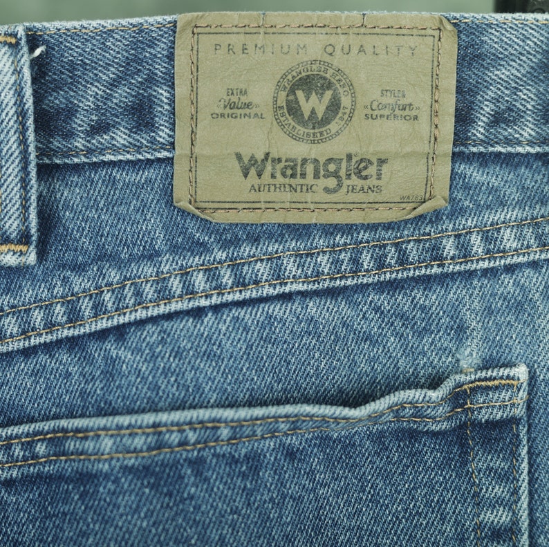 Peut inclure: Gros plan sur une poche de jean bleu denim avec un patch en cuir brun qui indique "Premium Quality" et "Wrangler Authentic Jeans".