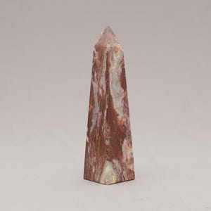 May include: A tall, obelisk-shaped stone sculpture. The stone has a reddish-brown and white marbled pattern. The sculpture tapers to a point at the top and has a square base. The object is standing upright against a plain background.