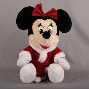 Pu&ograve; includere: Una bambola di peluche di Minnie Mouse, seduta e che indossa un abito rosso e bianco in stile Babbo Natale con un fiocco abbinato. La bambola ha orecchie nere, un viso color crema e un fiocco rosso. L'abito include una fascia rossa.