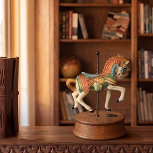 May include: A carousel horse figurine mounted on a central metal pole and a circular wooden base. The horse features a cream-colored body with a detailed saddle in shades of orange, green, and blue, and a textured mane with intricate patterns.