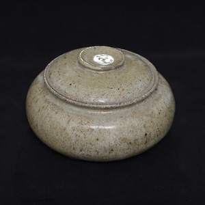May include: A round, low-profile ceramic lidded jar with a speckled, light beige and tan glaze. The container features a smooth, rounded body and a matching circular lid with a small, centered knob handle, showcasing a natural, earthy texture.