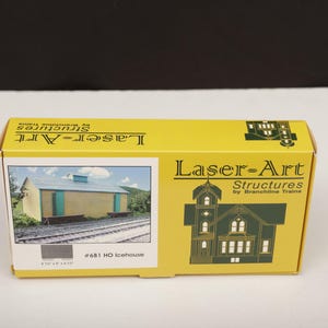 May include: A yellow box for a model train structure. The box features the text "Laser-Art Structures" and a picture of a building with teal doors and a blue roof. The box is labeled "#681 HO Icehouse".