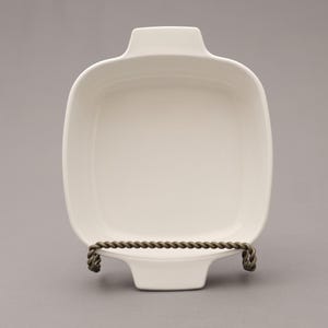 May include: A square, off-white ceramic baking dish with rounded corners and two handles. The dish is displayed on a decorative metal stand with a twisted rope design. The dish is empty and ready for use.