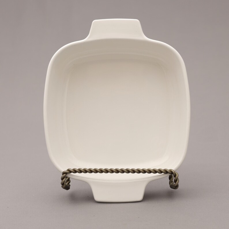 May include: A square, off-white ceramic baking dish with rounded corners and two handles. The dish is displayed on a decorative metal stand with a twisted rope design. The dish is empty and ready for use.