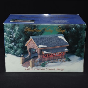 May include: A rectangular product box for a Heartland Valley Village Deluxe Porcelain Covered Bridge. The box features a graphic of a miniature covered bridge with a dark shingled roof, stone pillars, and red accents, set in a snowy landscape.