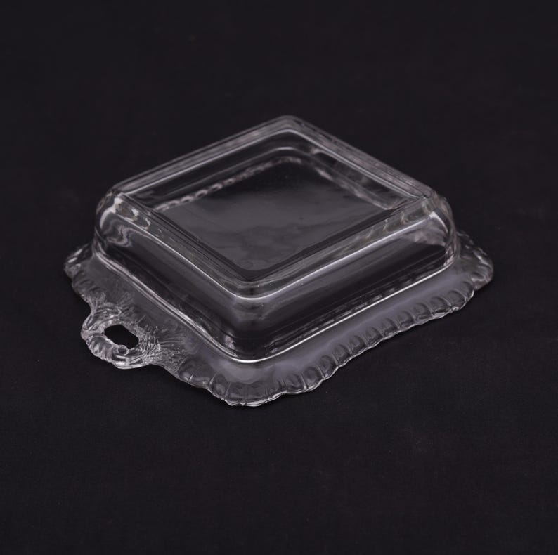 May include: A clear glass square dish with a lid. The dish has a decorative scalloped edge and a handle on each side.