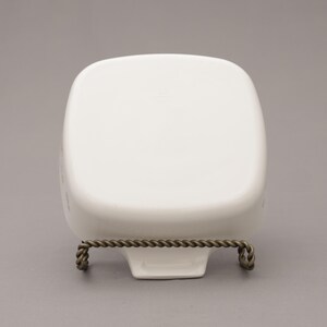 May include: A white, square-shaped ceramic dish with rounded corners, displayed on a decorative metal stand. The stand has a twisted rope design and is made of a dark, possibly bronze-colored metal. The dish is empty and sits on a gray background.