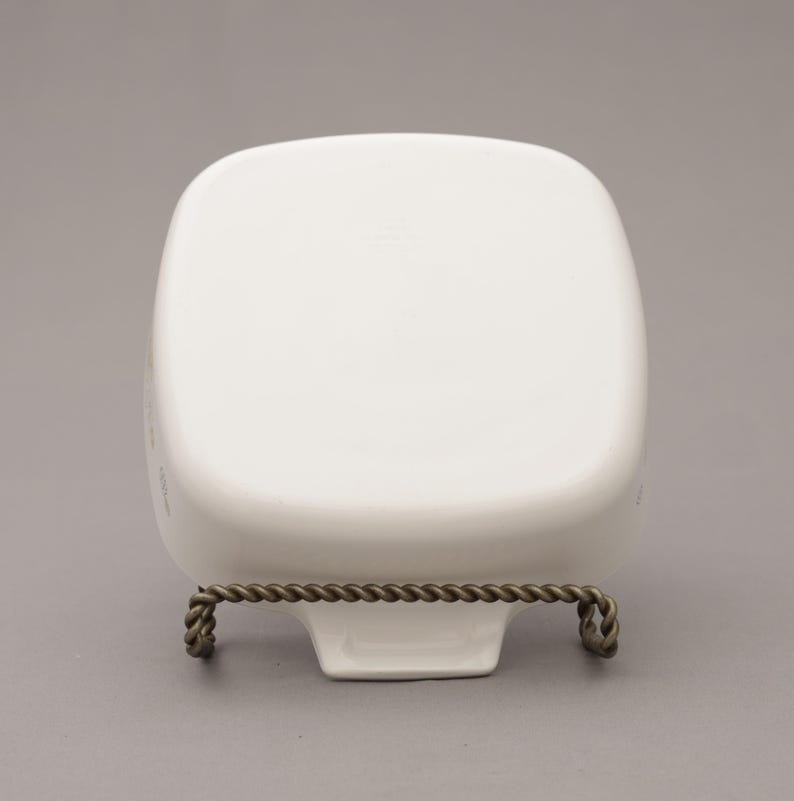May include: A white, square-shaped ceramic dish with rounded corners, displayed on a decorative metal stand. The stand has a twisted rope design and is made of a dark, possibly bronze-colored metal. The dish is empty and sits on a gray background.