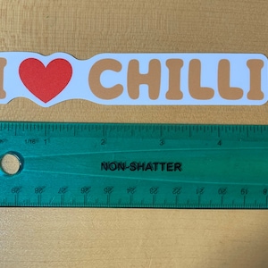 May include: A white sticker with the text "I ♥ CHILLI" in brown letters. A red heart is between the "I" and the "CHILLI". The sticker is measured against a green ruler.