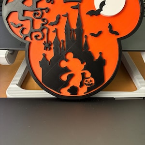 May include: A layered wall decoration shaped like a mouse head silhouette with a bright orange background. It features a black castle, a bare tree, a spider, bats, a white moon, and a small figure holding a jack-o'-lantern pumpkin.