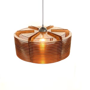 Puede incluir: Una moderna l&aacute;mpara colgante circular, hecha de madera marr&oacute;n claro en capas. El dise&ntilde;o presenta una serie de listones de madera horizontales alrededor de la circunferencia, con una secci&oacute;n superior decorativa. Una sola bombilla es visible en el interior, emitiendo un brillo c&aacute;lido.