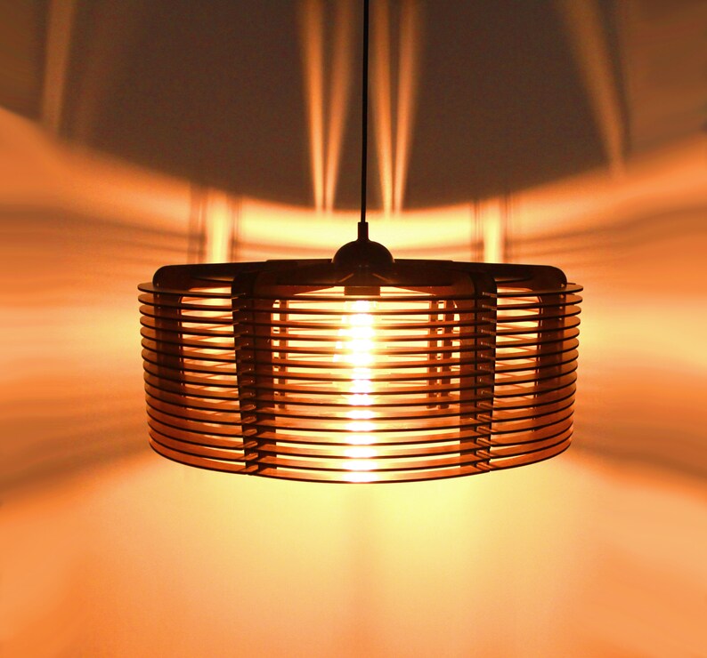 Puede incluir: Una moderna l&aacute;mpara colgante cil&iacute;ndrica hecha de listones de madera en capas. La c&aacute;lida luz de la bombilla interior proyecta un brillo suave y crea interesantes patrones de sombra en la pared. La l&aacute;mpara cuelga de un cable negro.