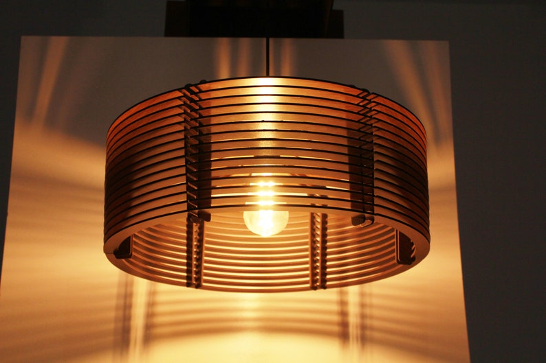 Puede incluir: Una l&aacute;mpara colgante cil&iacute;ndrica con un brillo c&aacute;lido y acogedor. La luz est&aacute; hecha de listones de madera en capas, creando un dise&ntilde;o &uacute;nico. La bombilla es visible en el centro, proyectando sombras en la pared. La l&aacute;mpara est&aacute; suspendida del techo.