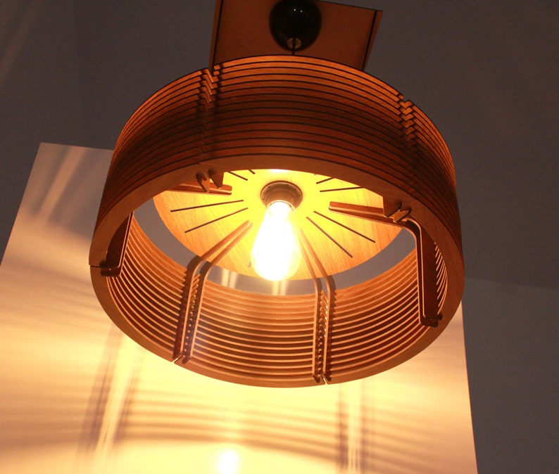 Puede incluir: Una l&aacute;mpara colgante de madera con una luz c&aacute;lida. El dise&ntilde;o circular presenta m&uacute;ltiples anillos de madera y una bombilla visible. La luz proyecta sombras interesantes sobre las superficies circundantes.