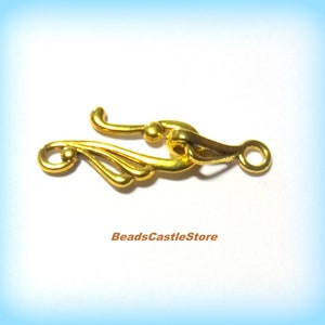 May include: Gold-tone metal clasp with a decorative, ornate design. The clasp has a loop on one end and a hook on the other.