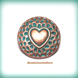 May include: A copper-colored button with a turquoise blue heart design. The heart is raised and has a textured surface. The button has a circular shape and a textured edge.