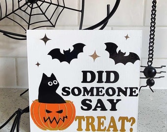 May include: A white wooden sign with a black cat sitting in a jack-o'-lantern. The sign says "Did someone say treat?" in black and gold letters. There are two black bats and gold stars on the sign.