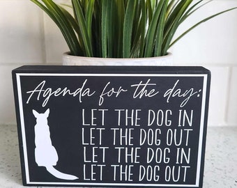 May include: Black wooden sign with white text that reads "Agenda for the day: LET THE DOG IN LET THE DOG OUT LET THE DOG IN LET THE DOG OUT". A white silhouette of a dog is in the upper left corner of the sign.