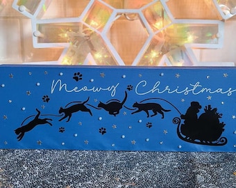 以下が含まれることがあります： 白い筆記体で「Meowy Christmas」と書かれた青い木製のブロック。黒い猫のシルエットが、中に人が乗ったそりを引っ張っています。足跡と星がブロック全体に散りばめられ、小さなキラキラとした装飾が施されています。