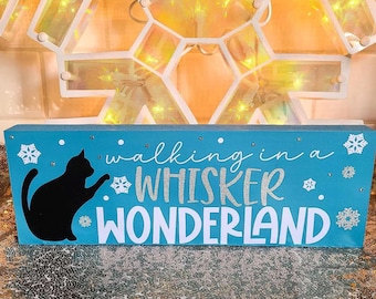 以下が含まれることがあります： 白い文字で「Walking in a Whisker Wonderland」と書かれたターコイズブルーのウッドサイン。黒いシルエットの猫が歩いている。サインはキラキラした仕上げで、白い雪の結晶で飾られている。