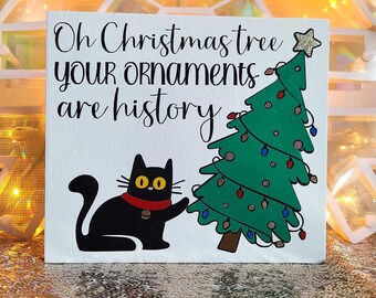 以下が含まれることがあります： 黒い猫と緑のクリスマスツリーが描かれた白い木製の看板。看板には「Oh Christmas tree your ornaments are history.」と書かれています。