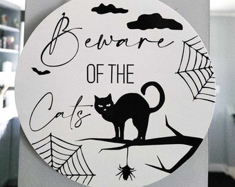 May include: A round, white sign with black lettering that reads "Beware of the Cats". The sign features a black cat silhouette, spiderwebs, and bats.