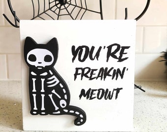 May include: White wooden block with a black and white skeleton cat illustration and the text "You're Freakin' Meowt".