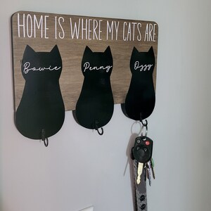May include: A wooden sign with the text "Home is where my cats are" and three black cat silhouettes with the names "Bowie", "Penny", and "Ozgy" written on them. Each cat silhouette has a hook for hanging keys.