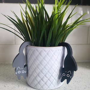 May include: A white ceramic planter with a diamond pattern, featuring two black cat-shaped plant markers. The cat markers are positioned on either side of the planter, with their tails extending upwards.