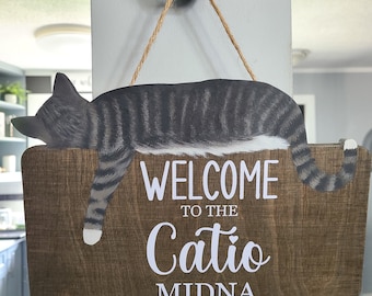 Puede incluir: Un letrero de madera con un gato rayado gris y blanco durmiendo encima. El letrero dice "Bienvenido al Catio Midna".